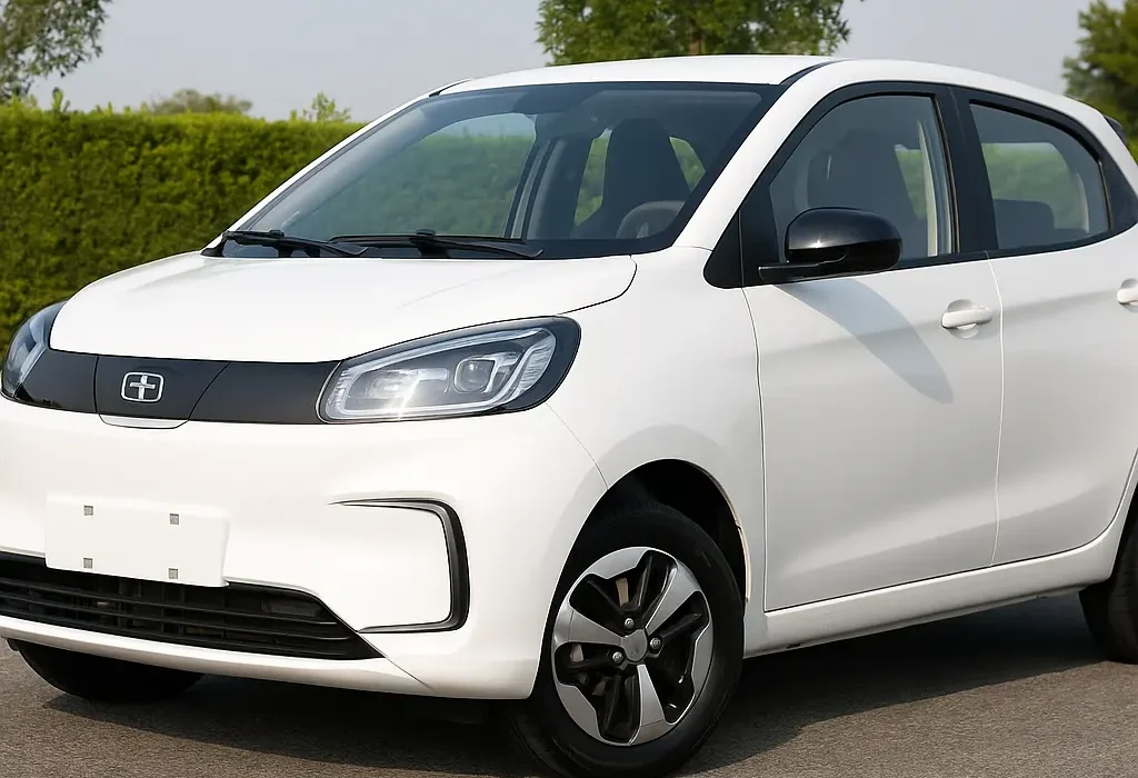 A digital photograph of a compact, white electric hatchback parked on a clean asphalt road with a backdrop of green hedges and tall trees under a bright, clear sky. The car features modern LED headlights, black alloy wheels, and a sleek, aerodynamic design, highlighting its eco-friendly and urban-focused appeal.